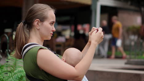 A-young-mother-with-her-baby-in-a-kangaroo-backpack-takes-photos-on-a-mobile-phone-while-traveling.-She-walks-and-glances-at-the-phone-screen-intermittently